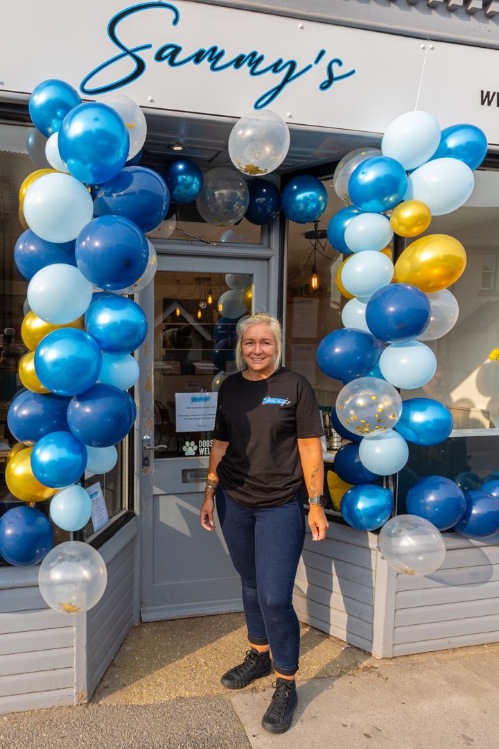 Sammy smiling in the doorway of the cafe, framed by an arch of blue and gold balloons